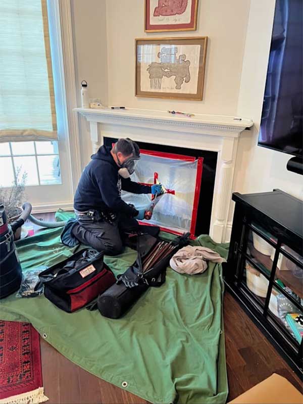 Stock photo of tech cleaning chimney wearing safety gear. Fireplace has plastic surrounding it, tools and vacuum on the floor with a tarp.